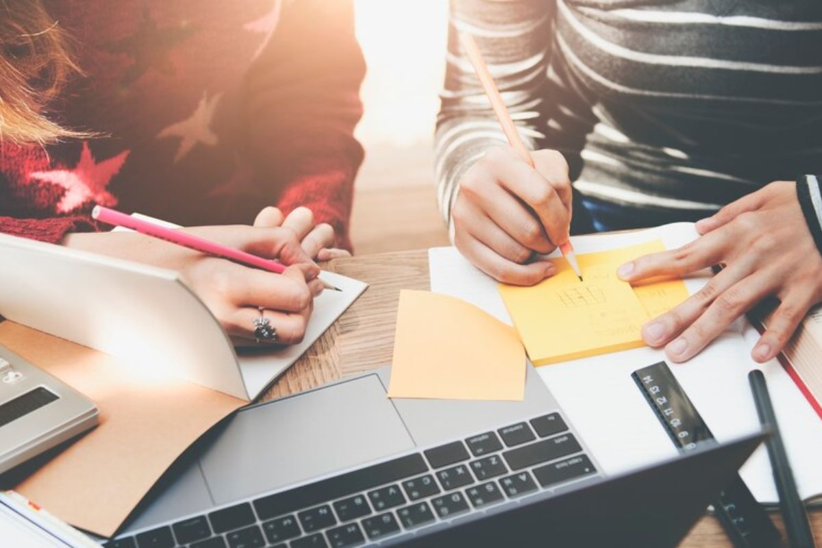 É possível ver dois estudantes sentado em uma mesa. Há diferentes materiais de estudos, inclusive um notebook