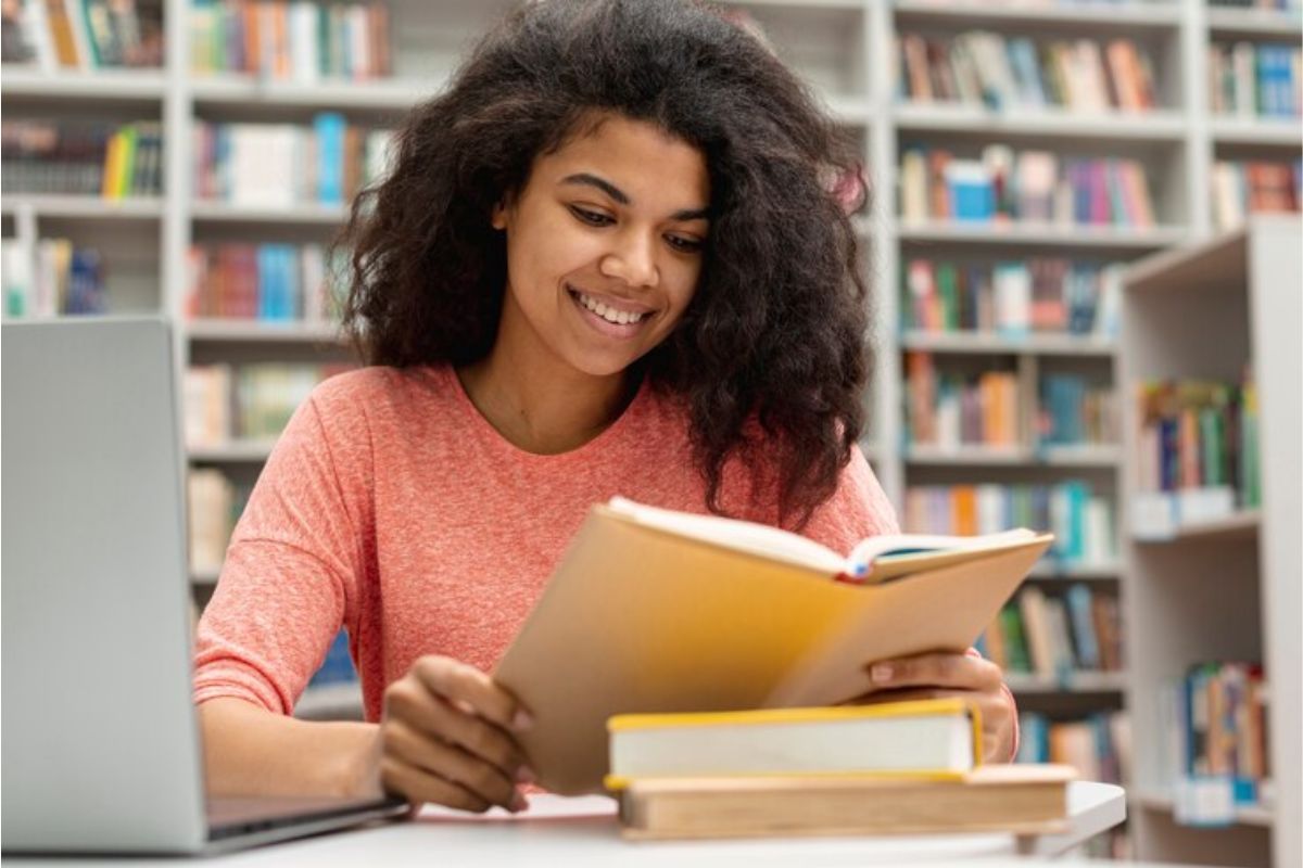 Uma estudante na biblioteca lê um livro. Ela está com o notebook aberto, onde participa da matrícula escolar MT 2025