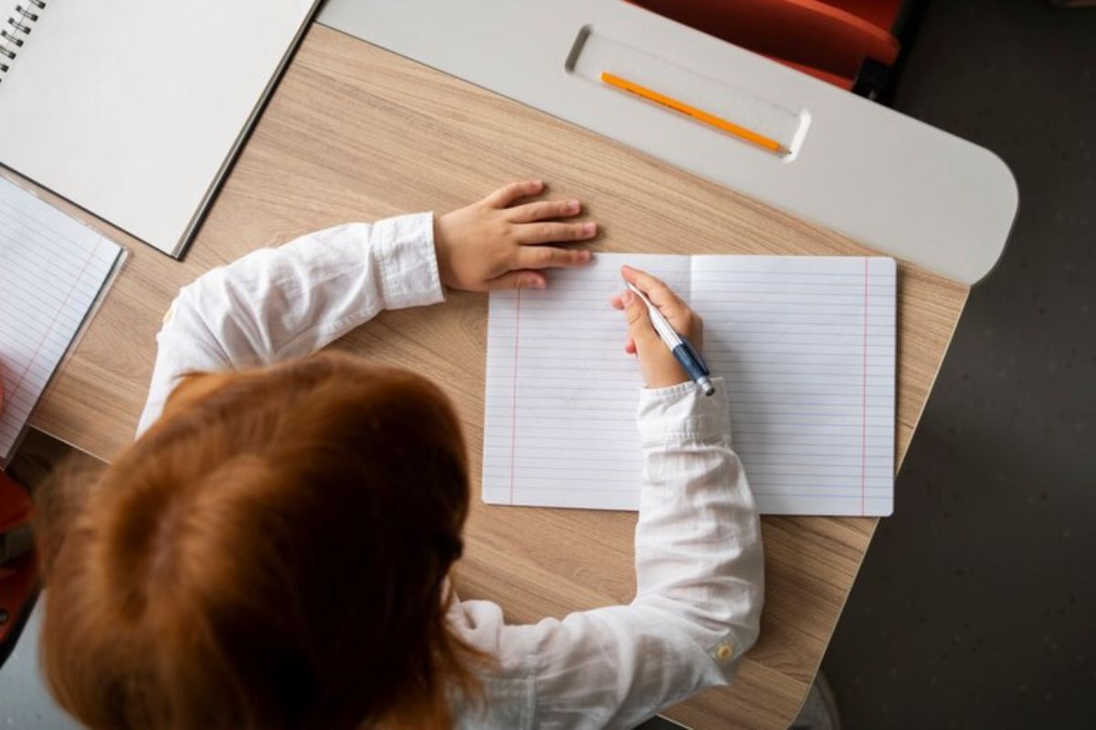 Uma aluna vista de cima, ela escreve em um caderno apoiada em uma mesa da sala de aula