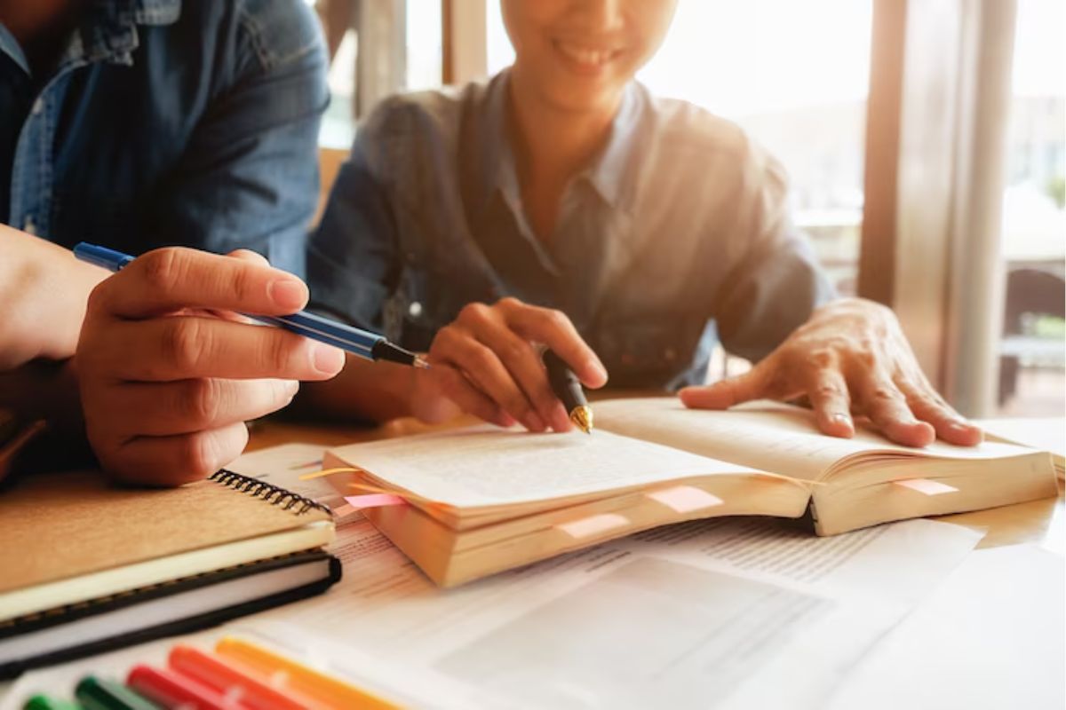 Dois alunos estudam juntos, eles estão com um livro aberto na mesa. Um estudante destaca algo apontando com a caneta