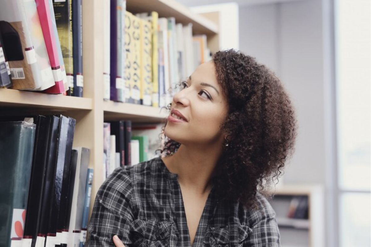 Uma estudante na biblioteca. Ela olha para uma estante de livros procurando algo pra ler