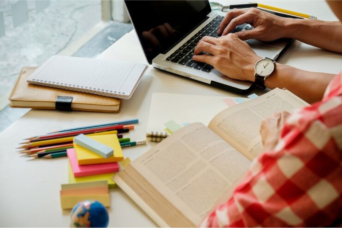 Dá para ver um jovem que mexe no notebook que está na mesa. Na mesa, há diversos materiais escolares que o ajudam no estudo.