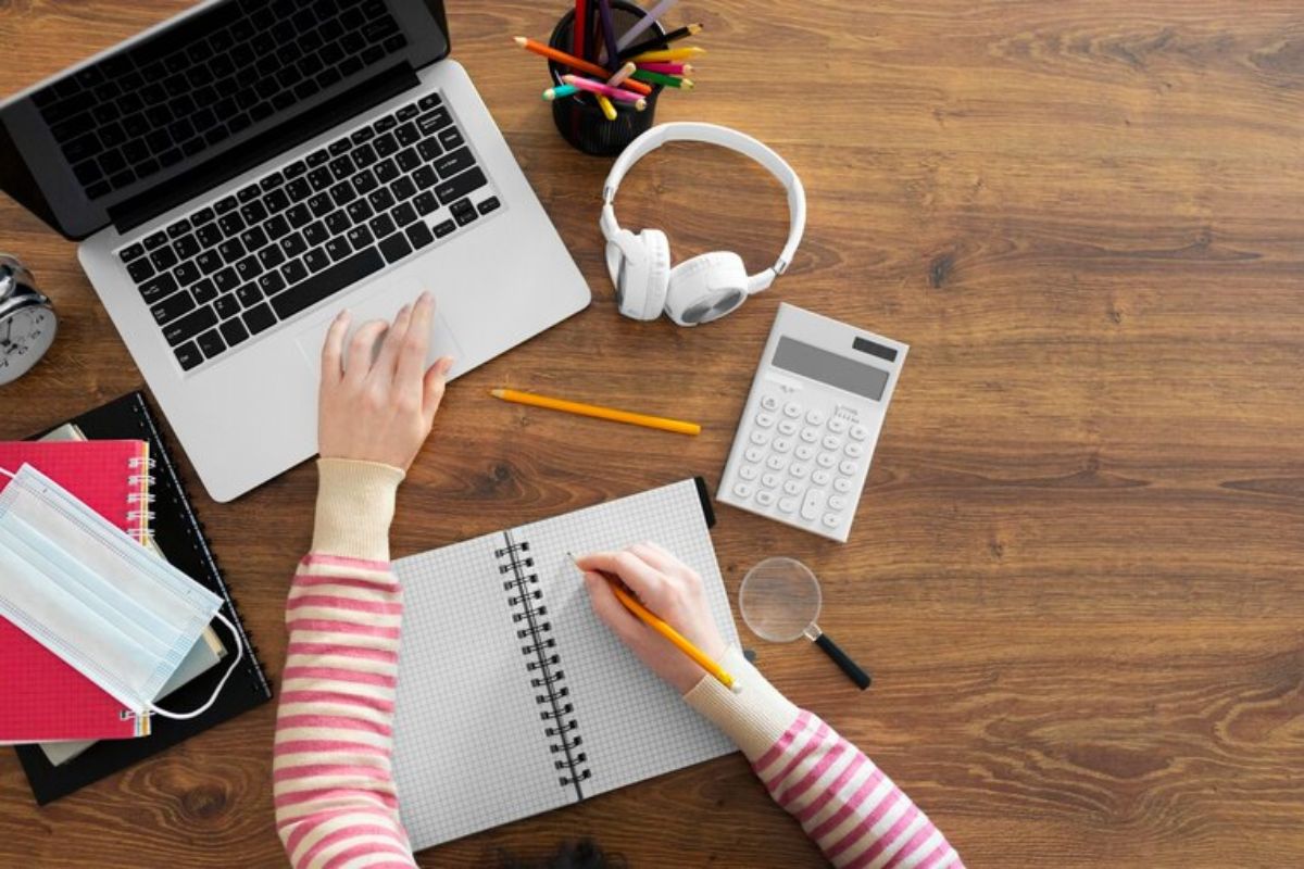 Vista de cima de uma aluna estudante com o notebook. É possível ver alguns materiais como um caderno aberto, uma calculadora, algumas canetinhas e cadernos fechados 