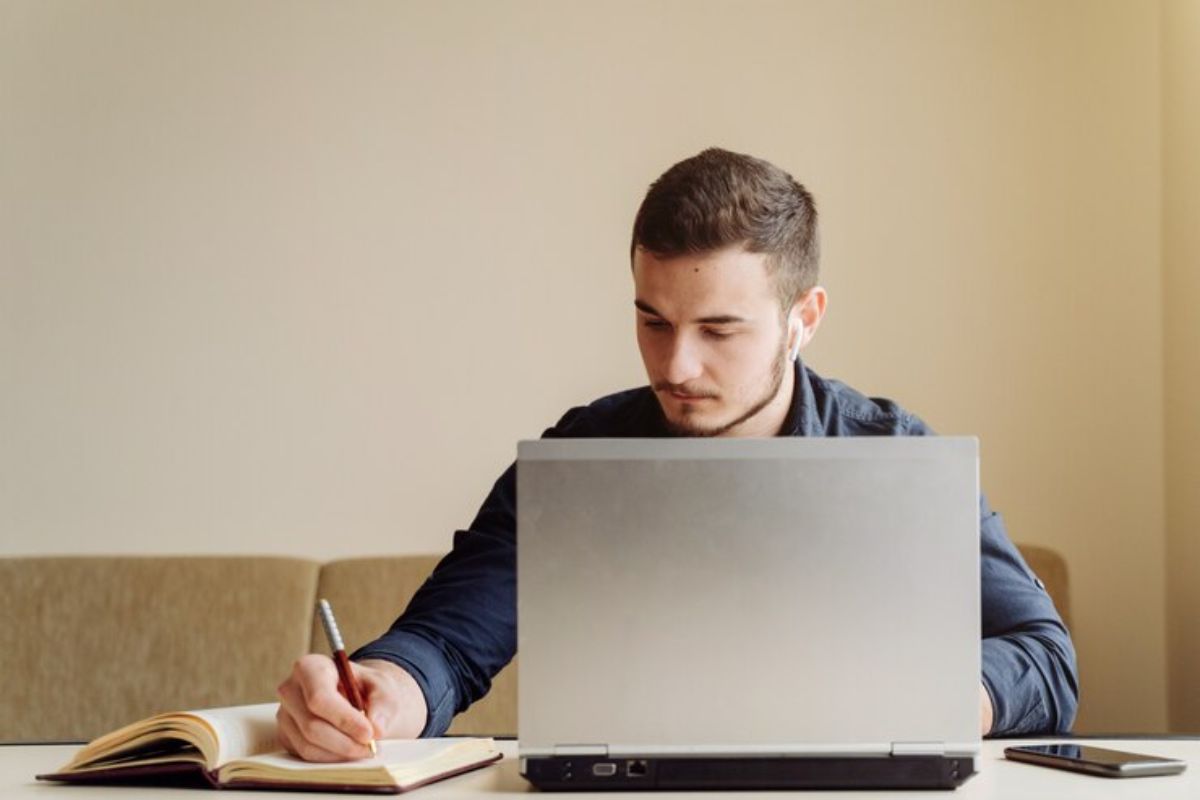 Um jovem estuda em frente ao seu notebook. Ao mesmo tempo ele escreve em seu caderno os detalhes sobre o vestibular UEA 2025