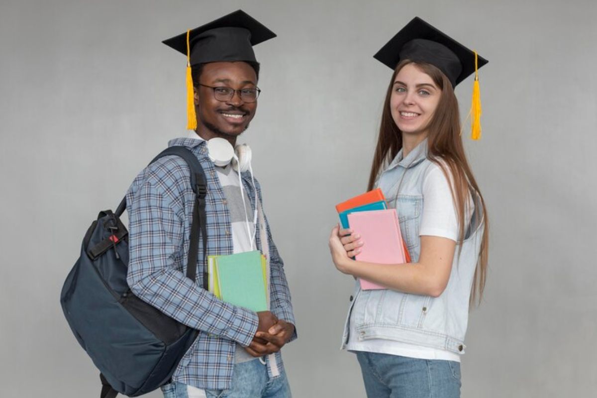 Dois estudantes estão segurando os seus materiais e sorriem para a foto. Há um jovem e uma menina 