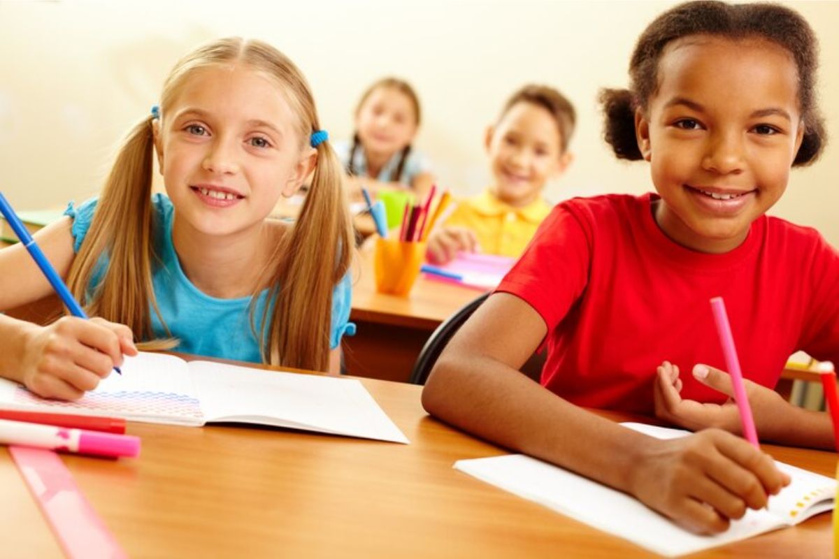 Quatro estudantes na sala de aula aparecem sorrindo para a foto enquanto escrevem em seus cadernos