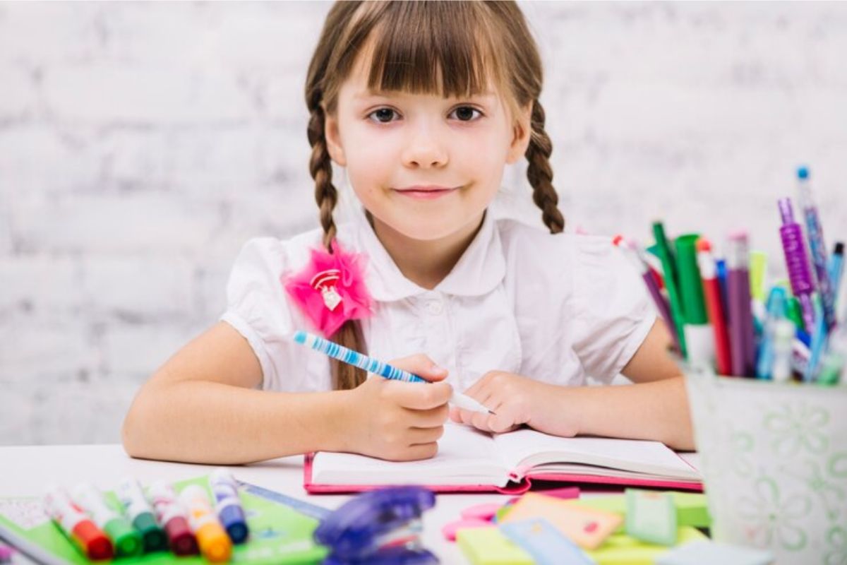 Uma menina escrevendo em seu caderno com diversos materiais escolares após sua mãe ver onde comprar material escolar barato em SP