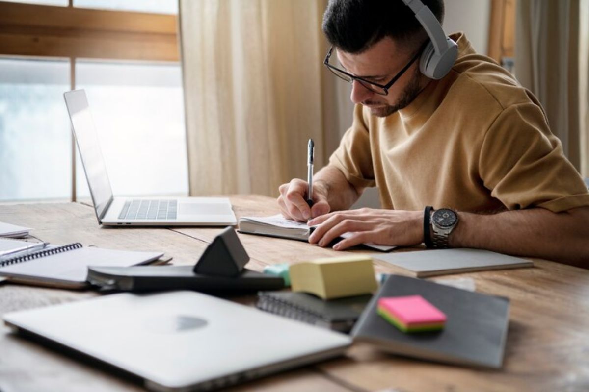 Um estudante em sua casa. Ele estuda em frente ao notebook, fazendo anotações em seu caderno sobre o CEFET-MG