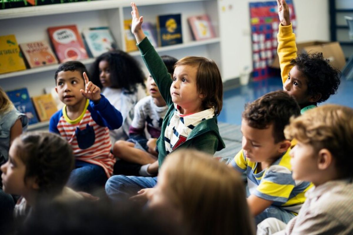 Uma turma de crianças sentadas no chão assistindo uma aula. Alguns alunos levantam a mão para fazer perguntas