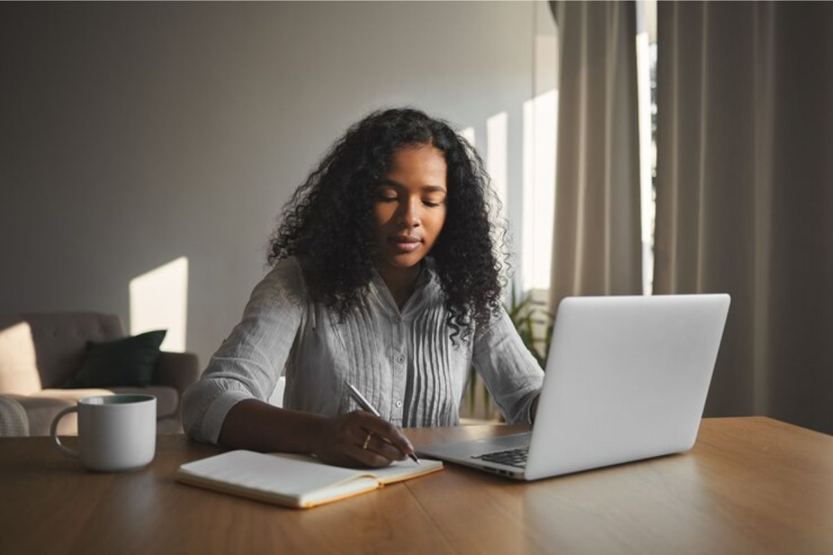 Uma estudante em frente ao seu notebook, ela participa da matrícula segundo semestre UNEB 2025. Ela faz anotações em seu caderno dos pontos mais importantes