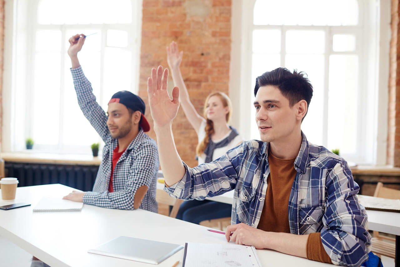 Jovens em sala de aula levantam a mão para responder questão do professor 